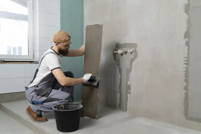 Muž nanáší lepidlo na dlaždici v koupelně během hydroizolace.