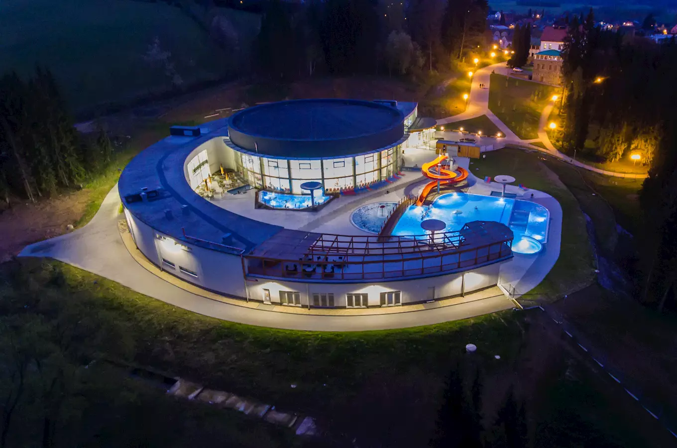 Nocní pohled na venkovní bazén s vodním hřištěm a krytým vnitřním prostorem.