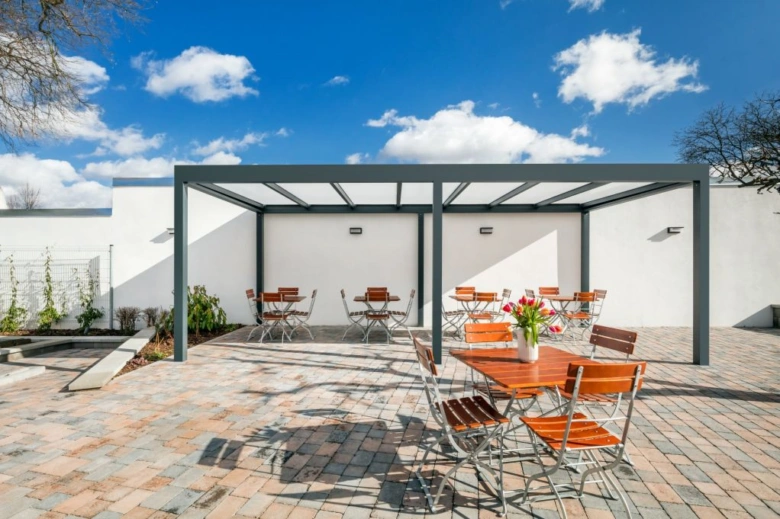 Moderní zahradní pergola s dřevěnými stoly a židlemi na dlažbě.