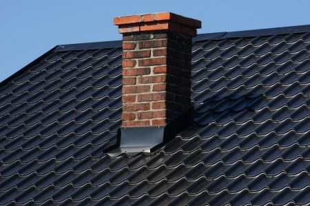 Cihlový komín na tmavé střechě s modrou oblohou.