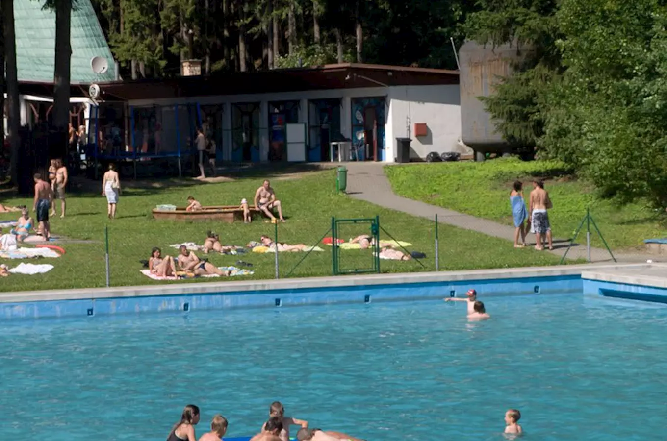 Venkovní bazén s lidmi plavajícími a opalující se na trávníku.