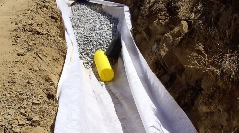 Bílá geotextilie s kamenivem a trubkami v příkopu pro drenáž.
