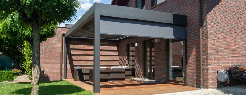Moderní hliníková pergola nad dřevěnou terasou u cihlového domu.