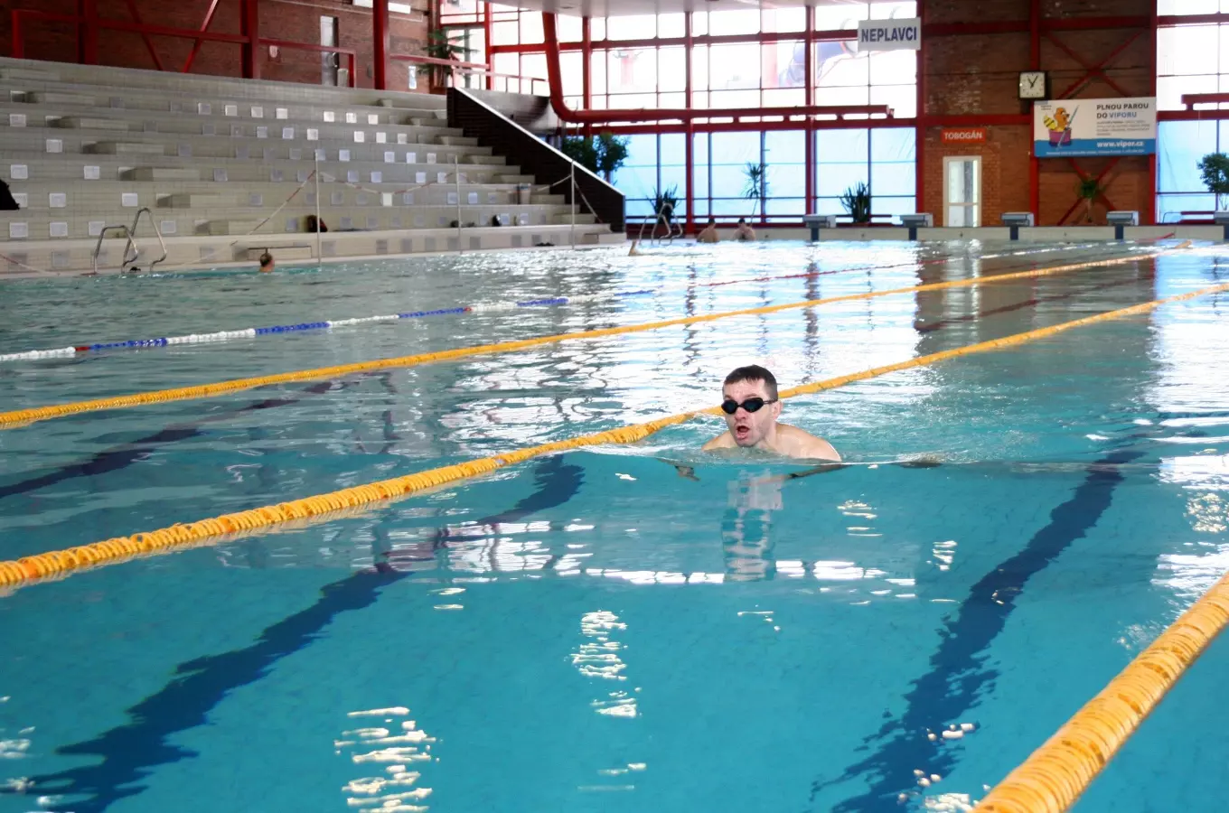 Muž plave na zádech v krytém bazénu s tribunami.