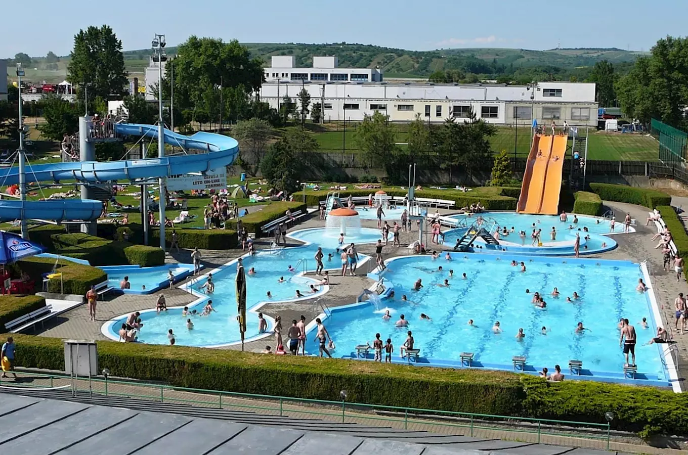 Venkovní aquapark s bazény a vodními skluzavkami plný lidí.