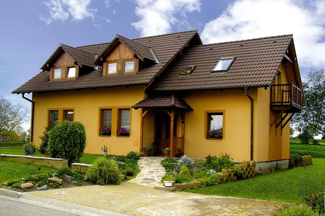 Žlutý dům s polovalbovou střechou na pozadí zelené trávy.