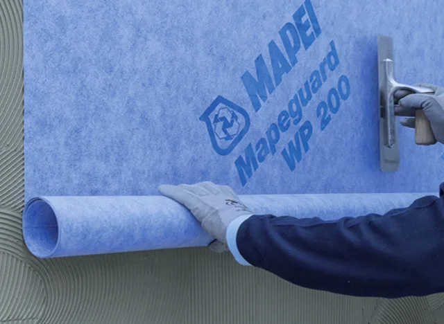 Pracovník aplikuje modrou hydroizolační fólii na stěnu v koupelně.