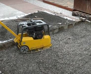 Žlutý vibrační lis na štěrkovém podkladu u betonového obrubníku.