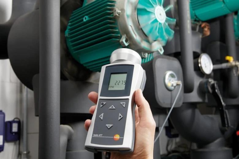 Ruční měřič rychlosti větru před průmyslovým ventilátorem.