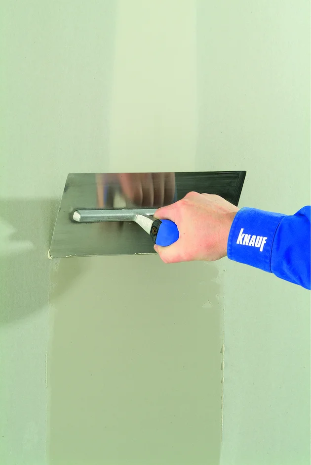 Ruka hladí štuk na zeď pomocí stěrky značky Knauf.