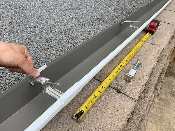 Montáž šedého okapového svodu s příchytkami a měřicí páskou.