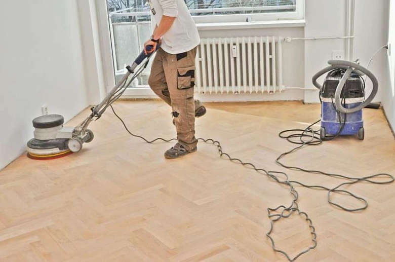 Muž brousí dřevěnou podlahu pomocí brusky a vysavače.