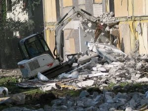 Bílý bagr rozebírá žlutou budovu s hromadou sutiny.