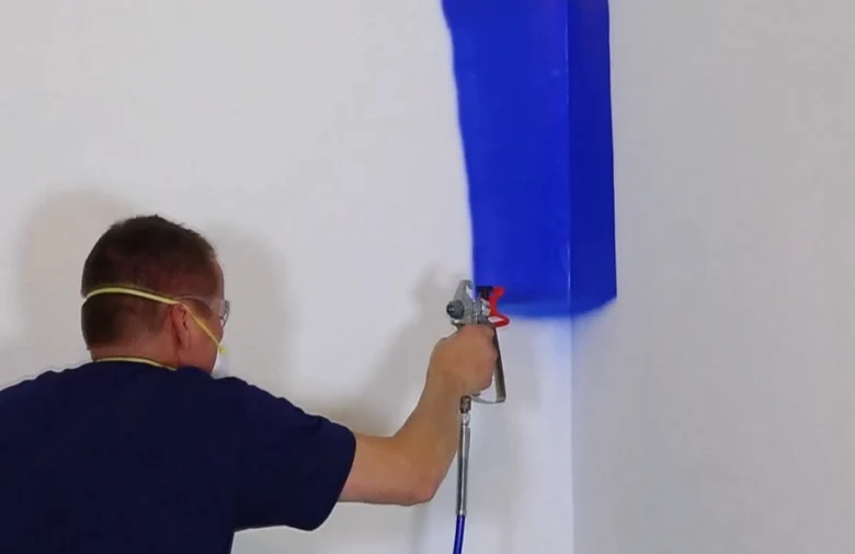 Muž stříká modrou barvu na bílou zeď pomocí stříkací pistole.