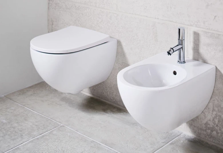 Bílá zdi montovaná WC sedacího křesla vedle bílého umyvadlového bidetu s chromovým kohoutkem.