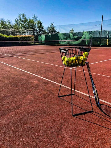 Tennis dvorek s košíkem míčů a raketou na slunci.